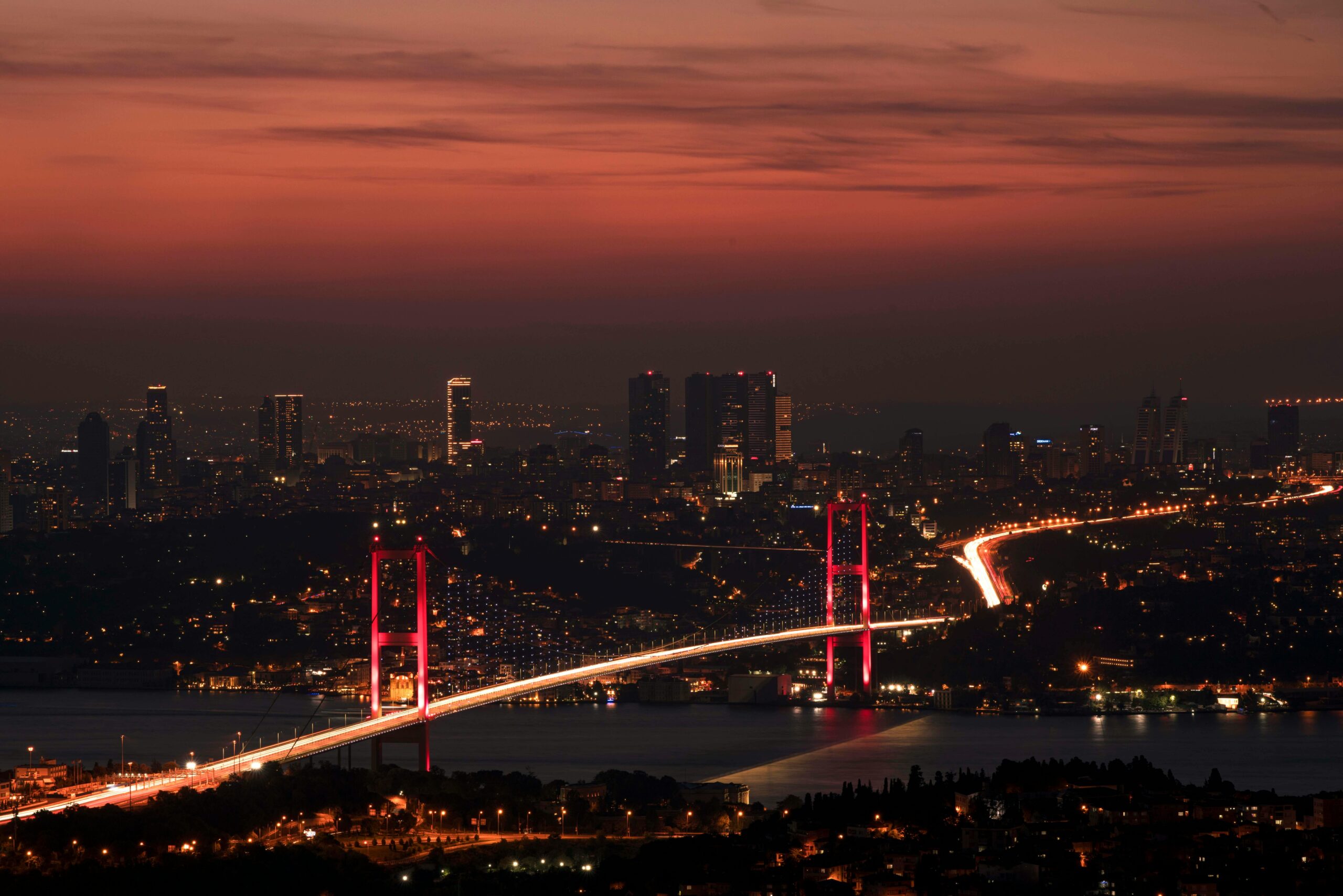 Home Stunning night view of the illuminated Bosphorus Bridge and Istanbul skyline under a red sky.
