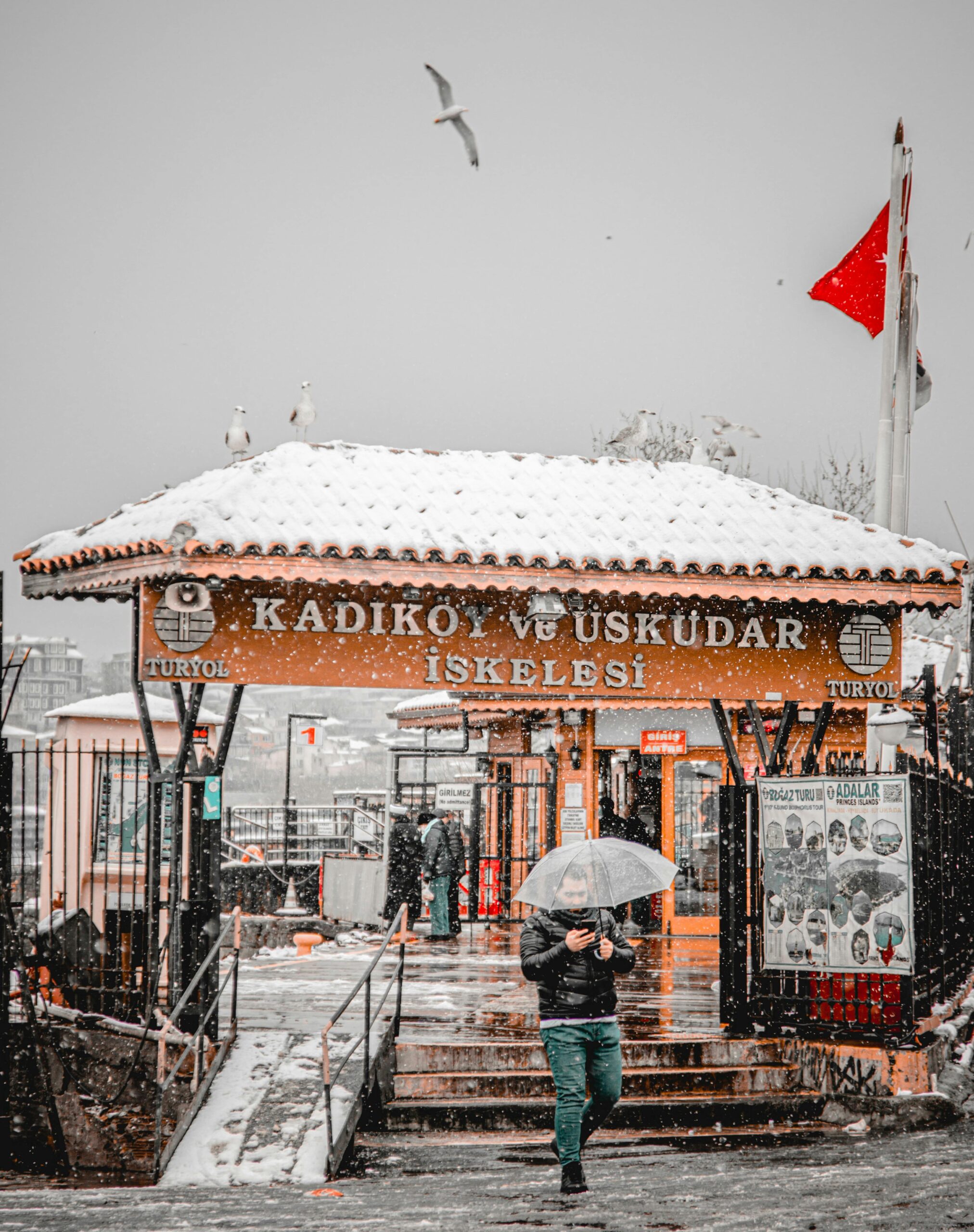 A person with an umbrella walks by Kadıköy Üsküdar Pier covered in snow, capturing a Turkish winter scene.